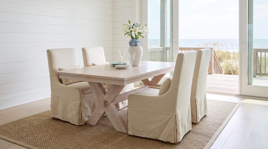 A whitewashed oak table on crossed trestle legs with a through-tenon, with four cream linen slipcovered host chairs, in an Isle of Palms beach house.