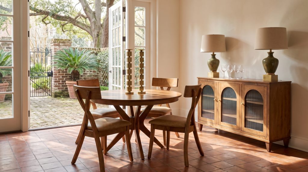 A round honey-finished table on a splayed pedestal, four chairs with crossing legs and tan leather seats, and a sideboard with arched reeded-glass doors.