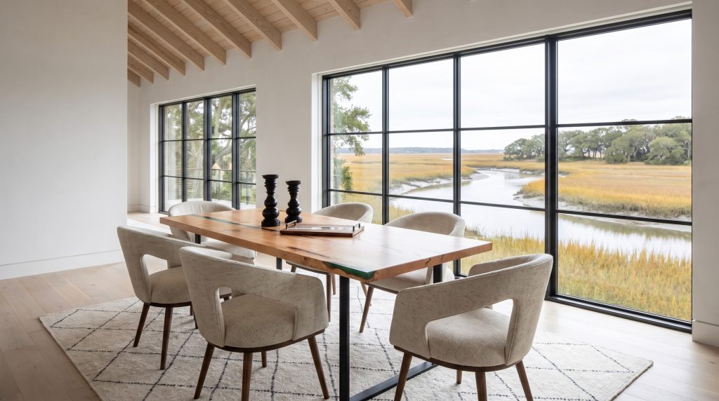 A pale maple dining table with a narrow emerald-green resin river and six oatmeal boucle open-back chairs in a Kiawah great room with glazing onto an autumn marsh.