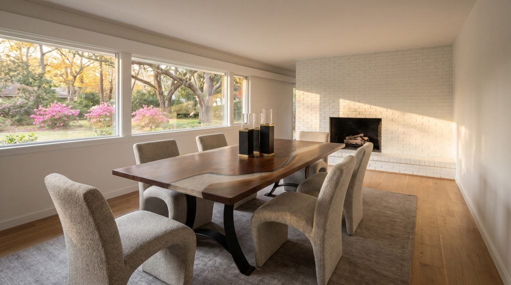 A thick walnut dining table with a pearl-silver resin river on arched black steel legs and six sculpted boucle chairs in a West Ashley ranch dining room with a white brick fireplace.