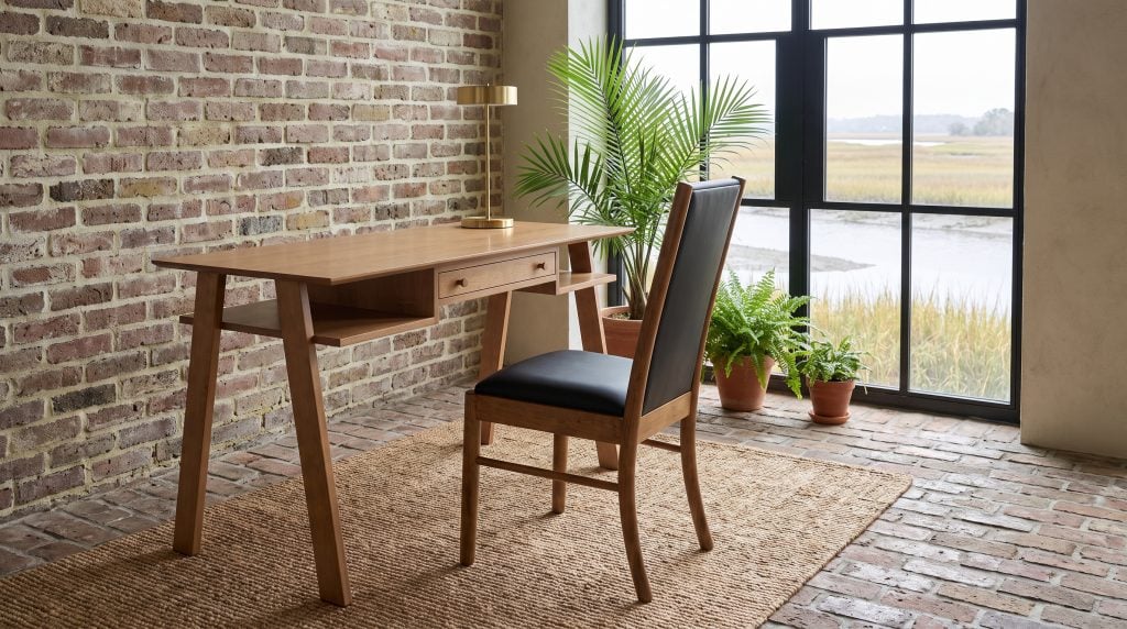 A honey-tan maple writing desk with outward-raking legs and a black leather side chair, in a brick sunroom over a tidal marsh