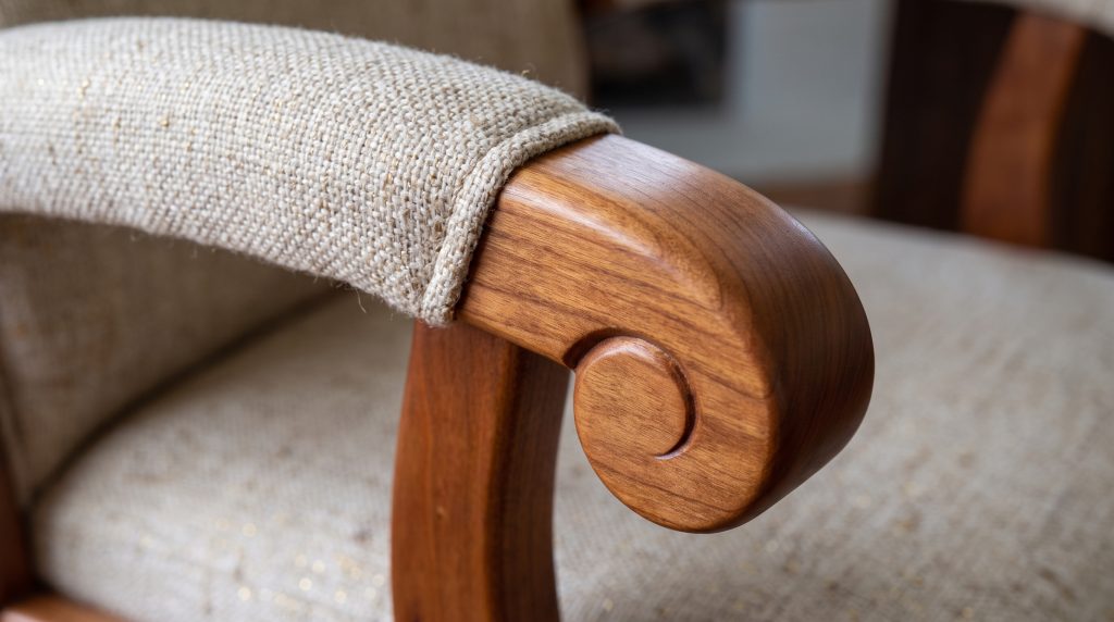 A close view of a coarse oatmeal tweed arm pad stitched down against the polished curve of a cherry scroll arm