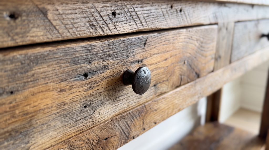 A close view of a small dark forged iron pull set into a reclaimed oak drawer front with saw marks and nail holes