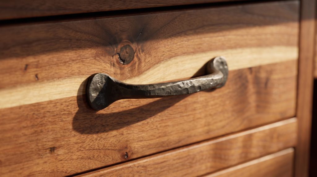 A close view of a hand-forged dark iron pull on an open-pored walnut drawer front with a pale sapwood streak