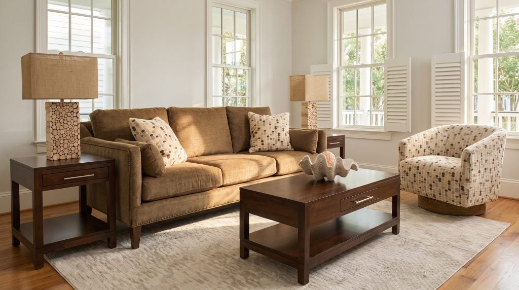 A tobacco-brown chenille sofa with brass nailhead trim, a dotted barrel swivel chair on a rope base and dark cherry tables, in a tall-windowed Mount Pleasant living room