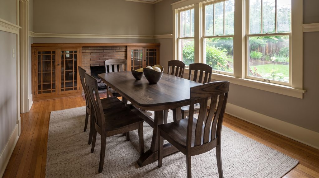 A quartersawn oak trestle dining table in a smoky shadow stain with six Oregon slat-back chairs, in a Craftsman bungalow dining room after rain