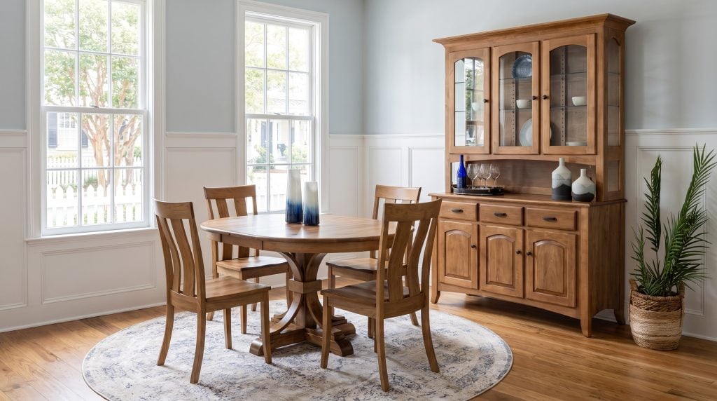 A rustic hickory round table with bowed-splat chairs and a lit maple china hutch, in an Old Village Mount Pleasant dining room on a bright morning.