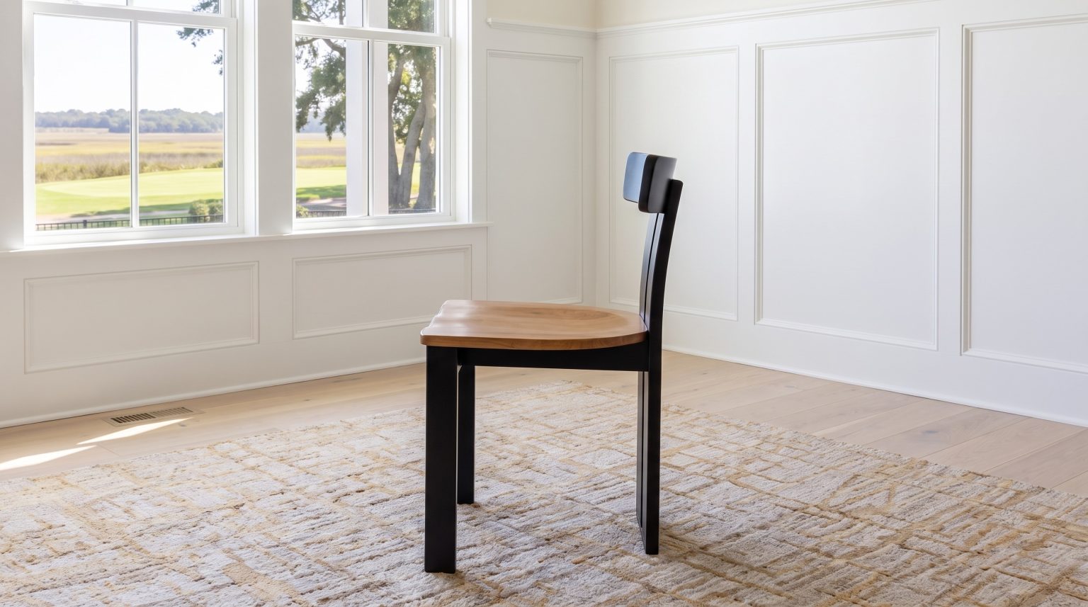 A single black-framed dining chair with a honey maple seat seen from the side on a sand rug