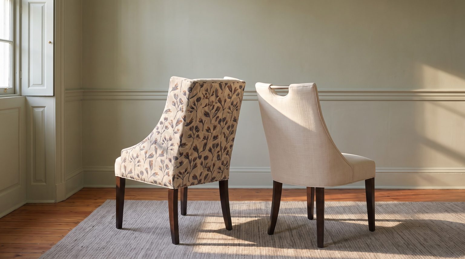 A floral-backed host chair beside an ivory linen side chair with a nickel pull, against sage paneling