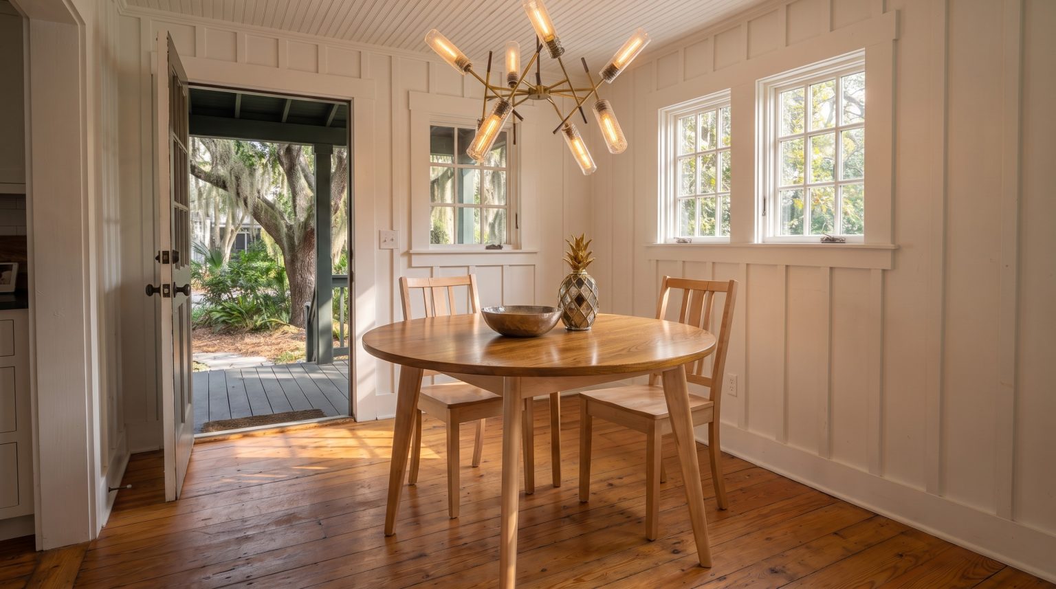 A round table with a golden elm top and pale maple legs, two chairs beyond, in a board-and-batten room