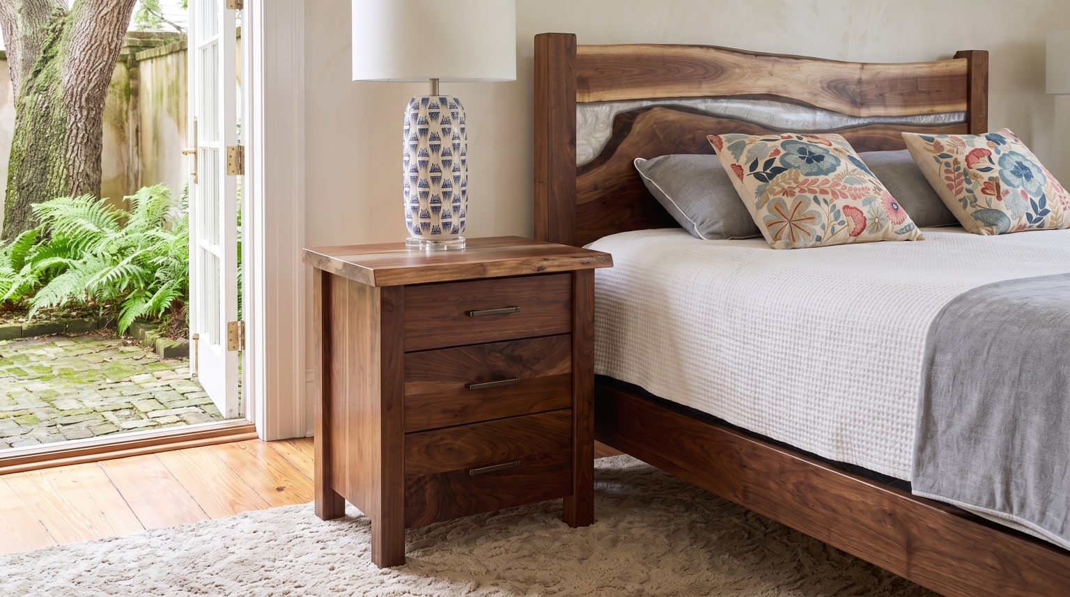 A walnut three-drawer nightstand with a live-edge top and a blue and white lamp beside a bed