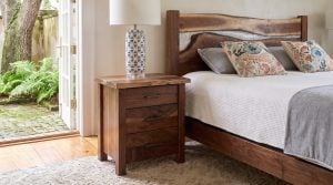 A walnut three-drawer nightstand with a live-edge top and a blue and white lamp beside a bed