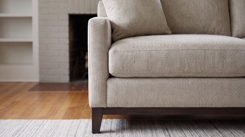 A close view of an oatmeal woven sofa arm and seat cushion on a dark wood base
