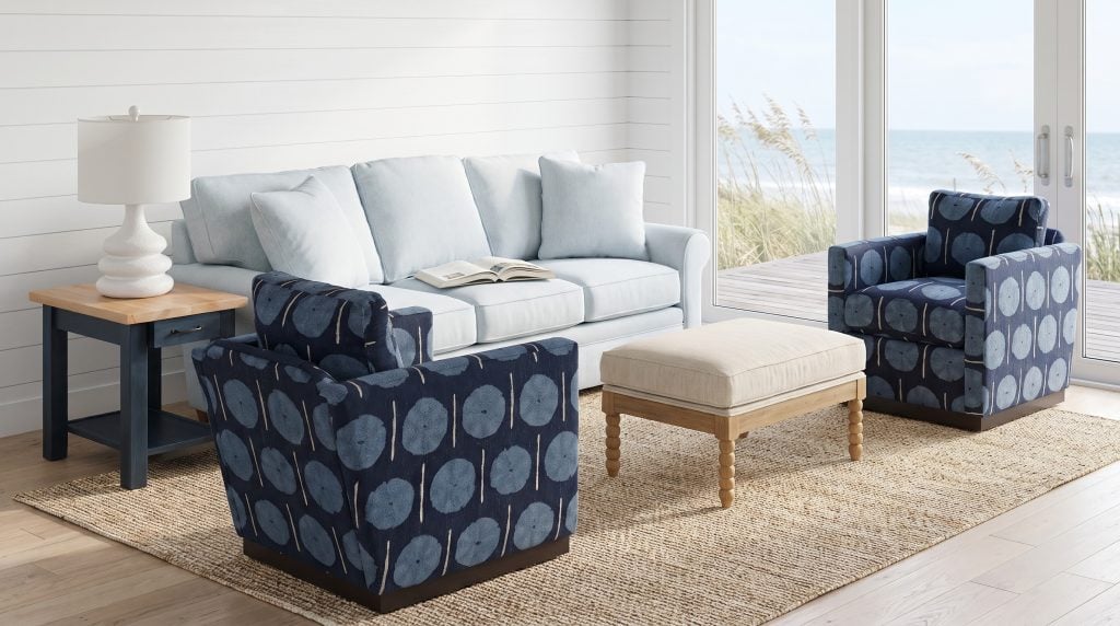 Two swivel chairs in a navy dandelion print, an ice-blue sofa, a spool-leg ottoman and an ink-blue end table in a white shiplap room open to the dunes