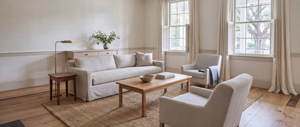 A Coastal Lowcountry living room: oyster linen sofa, quarter-sawn white oak coffee table and a woven seagrass rug, with late afternoon light coming through half-open louvered shutters.