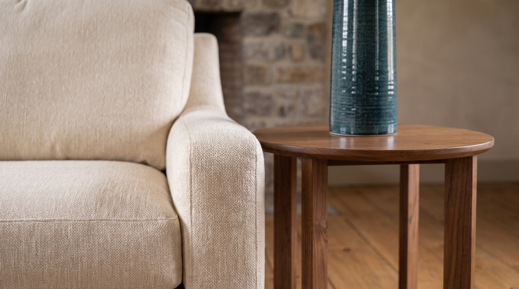 A cream woven sofa arm beside the edge of a round walnut end table and a teal ceramic lamp against dry-stacked stone