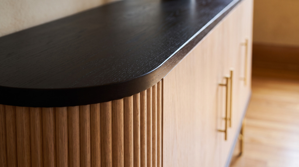 A close view of the fluted white oak buffet front, its black rolled top edge and pale gold pull catching low afternoon light