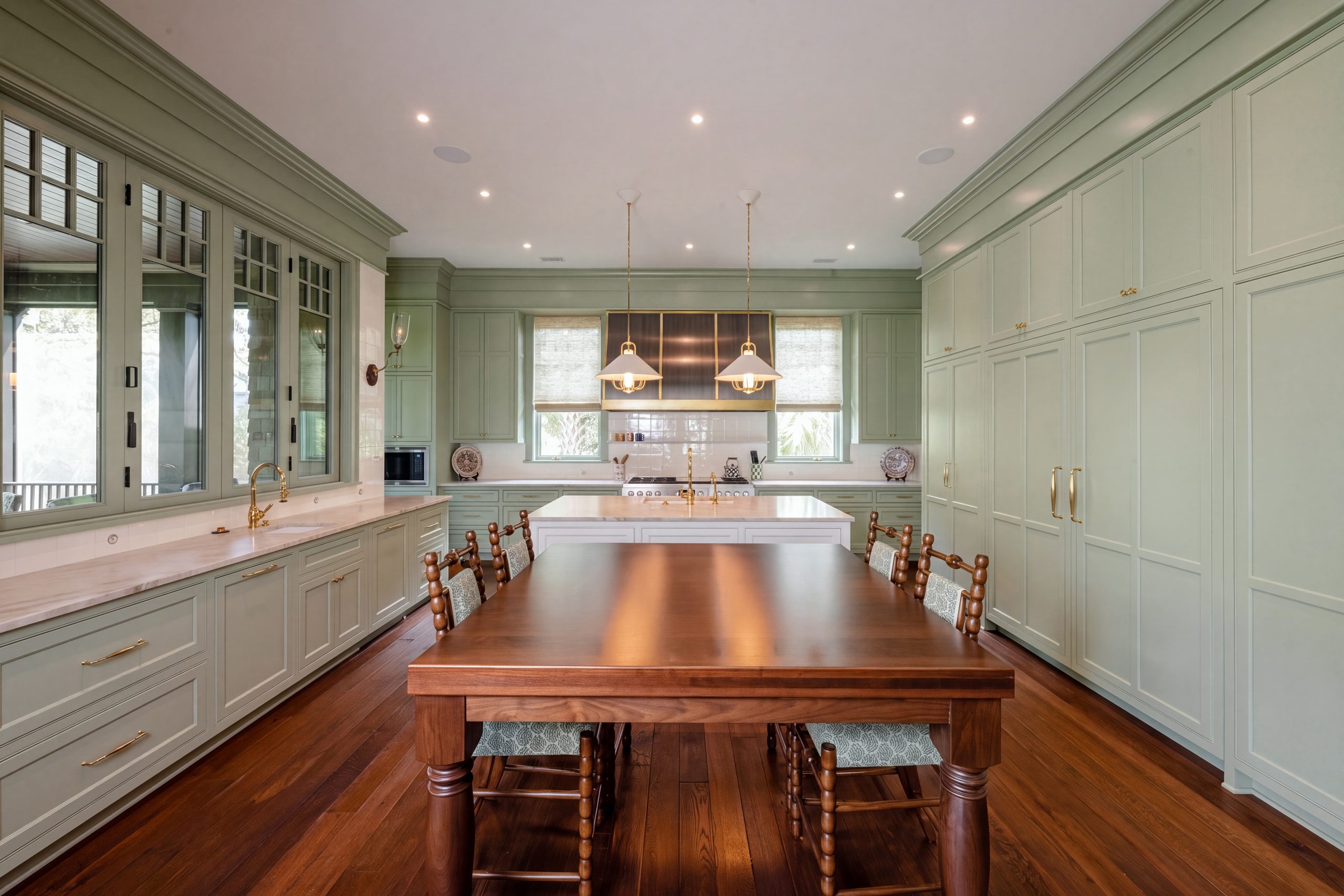 A long Kiawah kitchen with sage-green inset cabinetry, a floor-to-ceiling pantry wall, a white island and a wood table with turned chairs.
