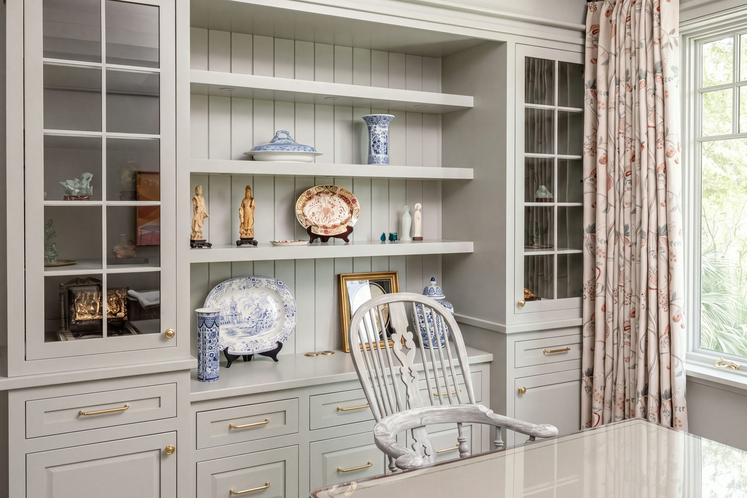 A painted built-in with mullioned glass doors, beadboard shelves of blue-and-white porcelain and brass-pulled drawers in a Kiawah Island study.