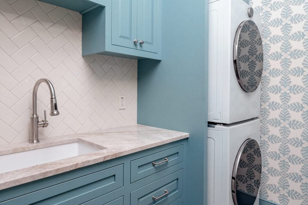A laundry room with blue raised-panel cabinetry, a quartzite counter and utility sink, herringbone tile and a stacked washer and dryer in a Kiawah Island house.