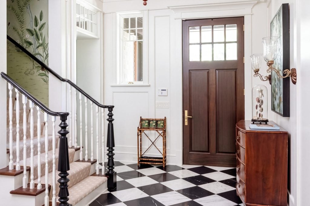 An entry hall with white panel millwork, a stair with white balusters and black newels, a black-and-white marble floor and a mahogany front door.