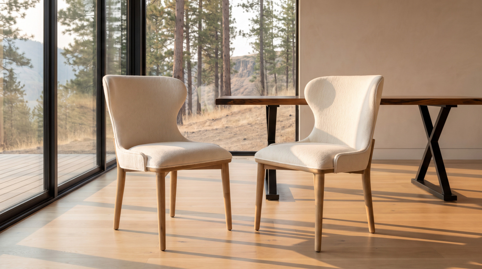 Two cream upholstered dining chairs with wooden legs backlit against a glass wall onto a pine treeline