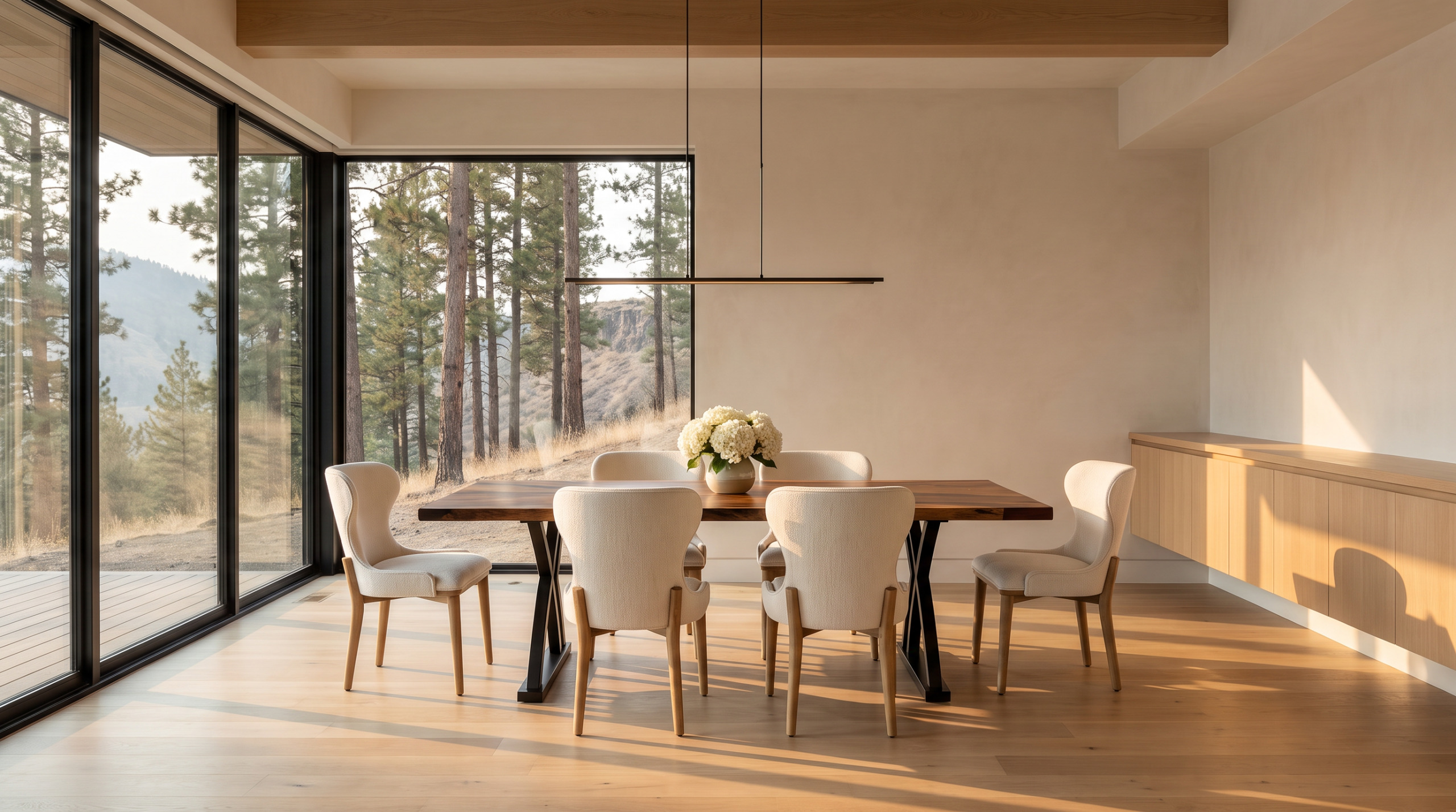 A live-edge walnut dining table on a black sculptural base with six cream upholstered chairs beside a glass wall onto pines