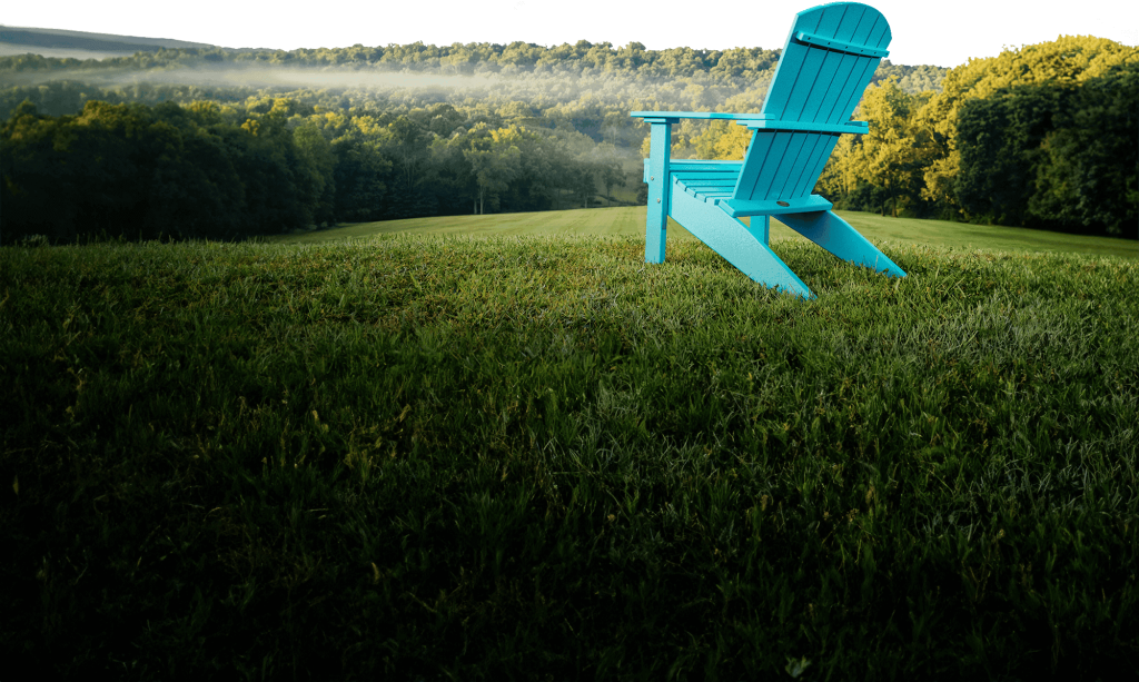 A single Aruba Blue poly Adirondack chair on a mown hillside at first light, mist over the treeline