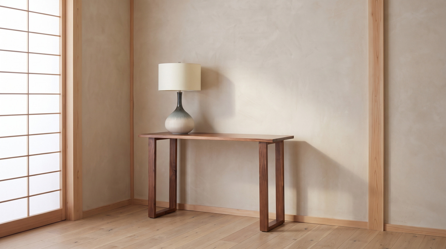 A slim walnut console table on open loop legs against a plaster wall with a ceramic table lamp on it