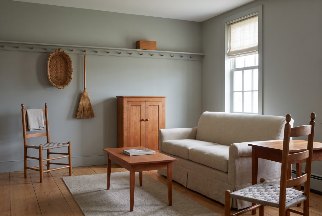 A Shaker living room: a cherry cabinet with wooden knobs, ladder-back chairs with woven tape seats, and a peg rail along a milk-painted wall.