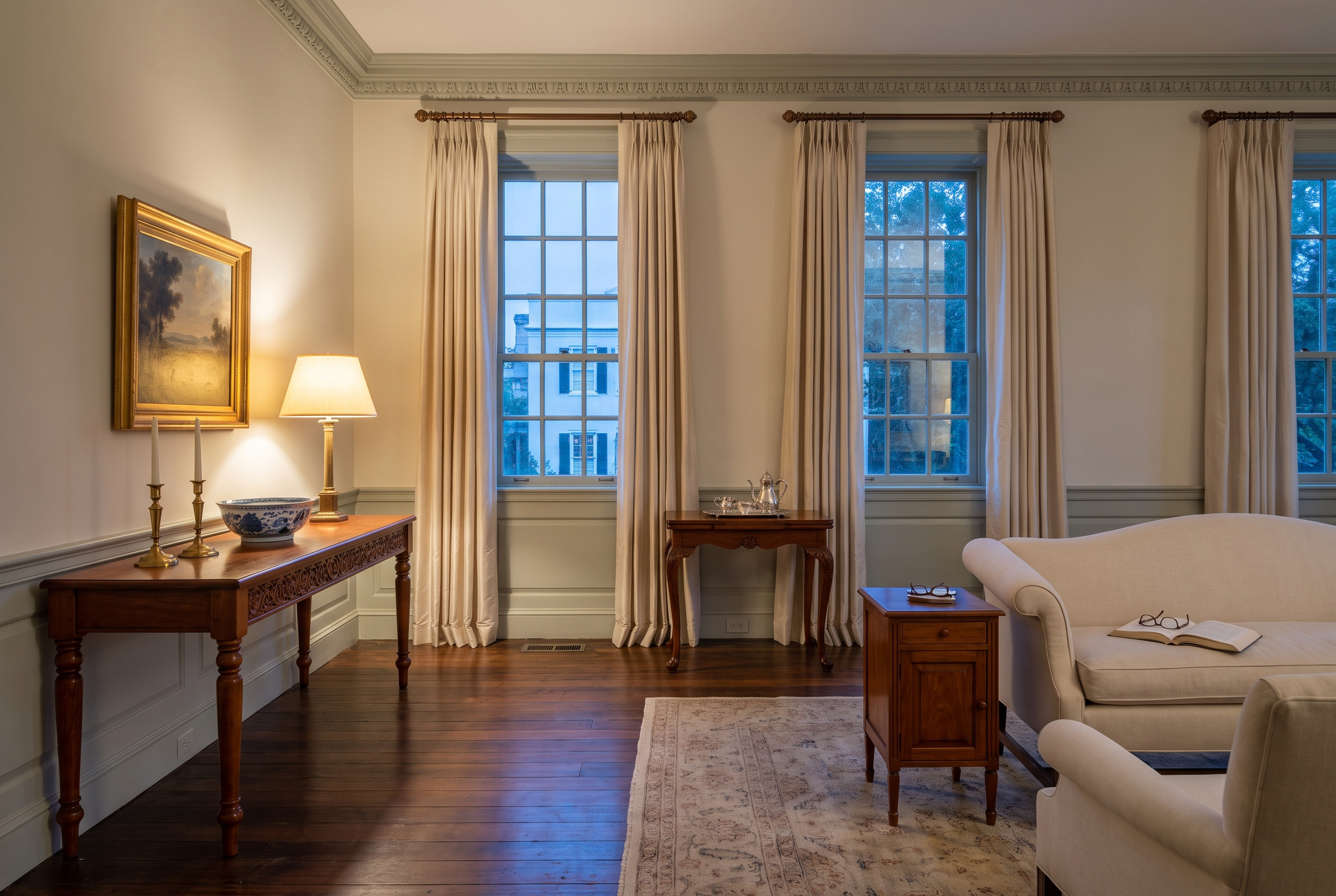 A formal Charleston drawing room: a carved cherry console with turned legs under a gilt landscape, a cherry tea table with cabriole legs, lamplit against blue evening windows.