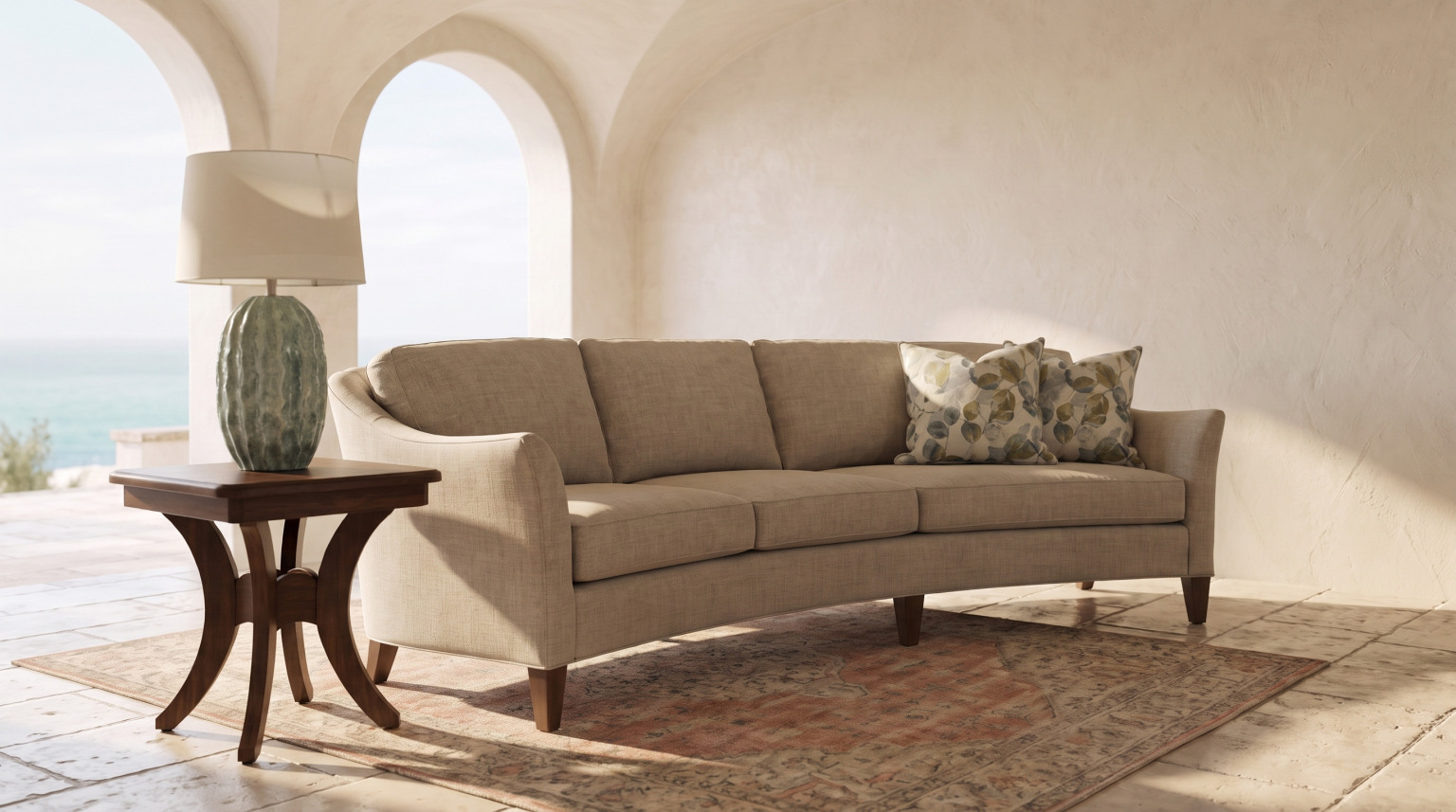 A curved-back sofa in a sand weave with a flared arm and tapered wood legs, beside a brown maple end table and lamp