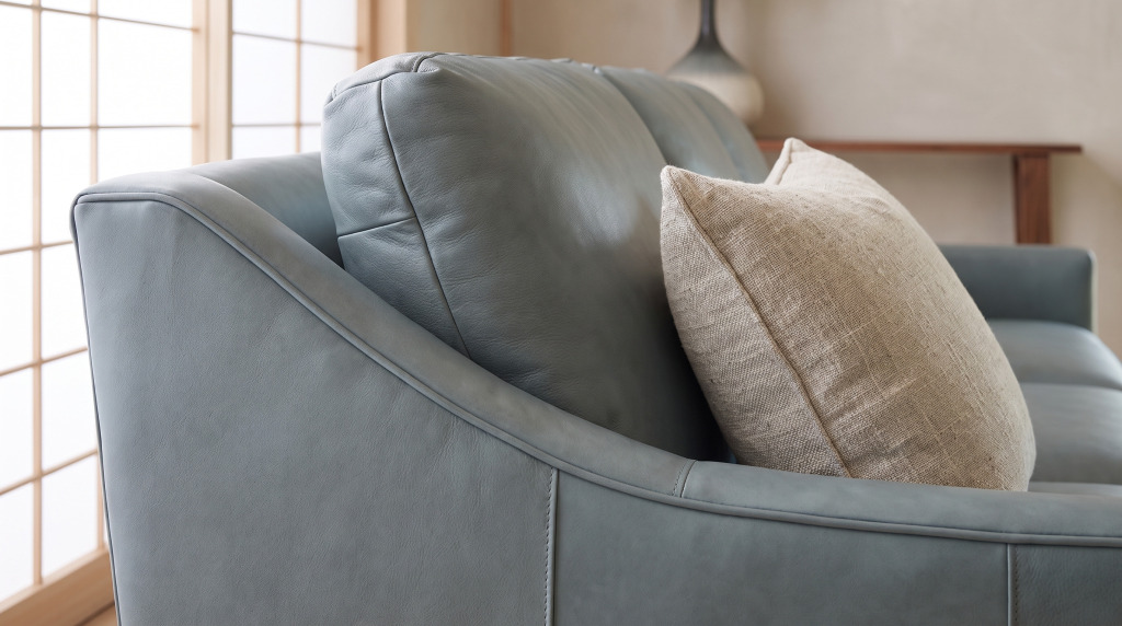 A close view of the rolled leather arm of a gray-green sofa with a welted seam and a linen cushion