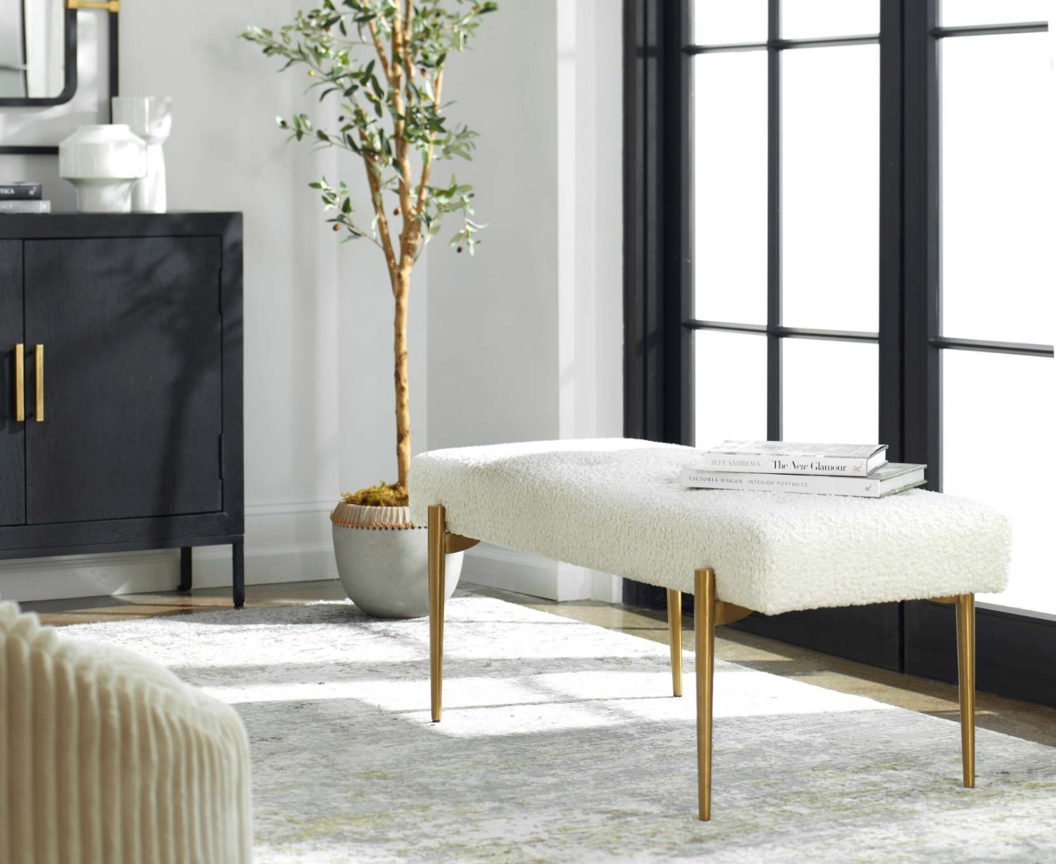 A boucle upholstered bench on slim tapered brass legs beside a dark cabinet
